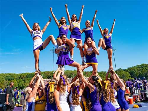 Thumbnail: Golden Rams cheerleaders in purple and gold uniforms forming a multi-level pyramid stunt on a sunny day at a football field, with clear blue sky overhead and a crowd visible in the background.