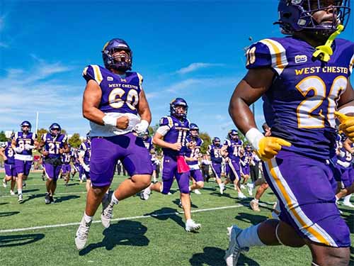 Thumbnail: Players from the West Chester University Golden Rams football team in purple and gold uniforms charging onto the field under a clear blue sky — filled with energy and motion as the squad enters the game together.