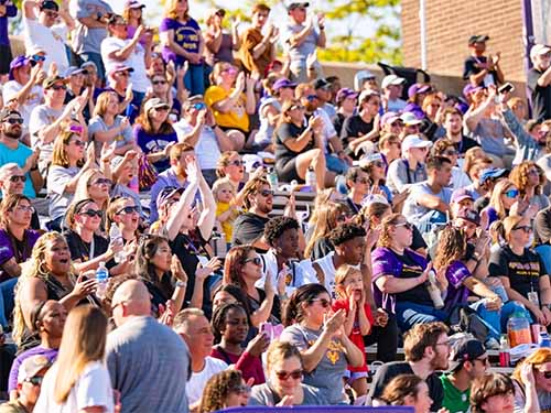 Thumbnail: Crowd of fans seated and standing in the bleachers at a West Chester University football game, many wearing purple and gold — cheering and waving their arms under daylight sky in support of the team.
