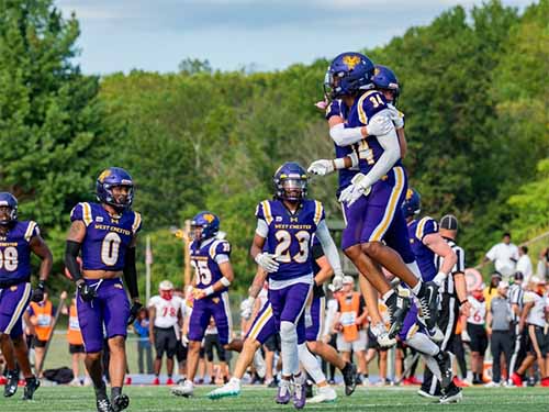 Thumbnail: Several members of the West Chester University Golden Rams football team in purple and gold uniforms on a sunlit field — two players mid-air in a celebratory jump, surrounded by teammates, with fans and sideline staff visible in the background.