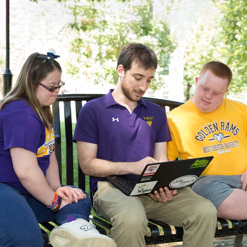 Three people sitting on a bench. The middle man is holding a laptop the woman and man (students) are looking at the laptop screen.