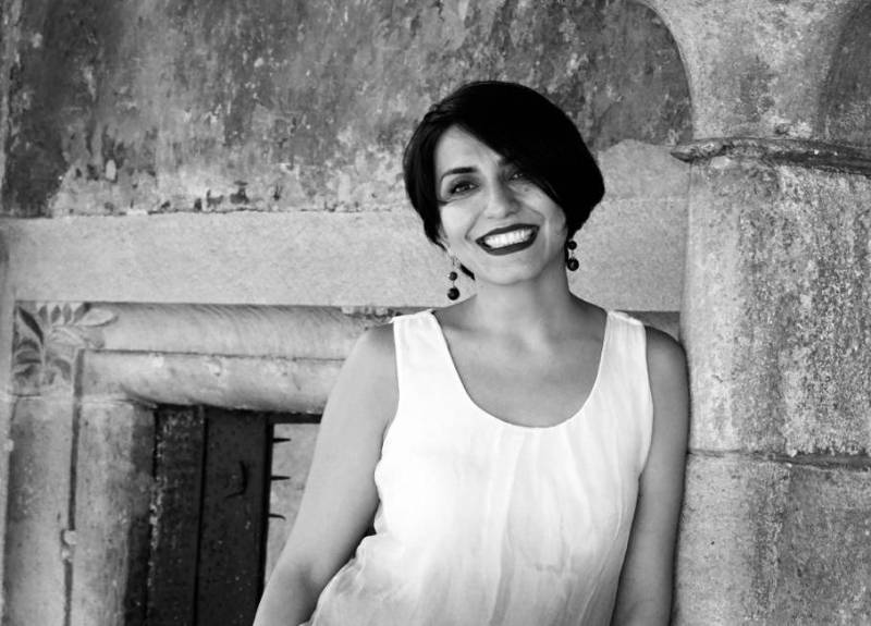 image of sara - feminine person with short dark hair, dark lipstick, hanging earings, a white sleeveless blouse with an architectural cement background 